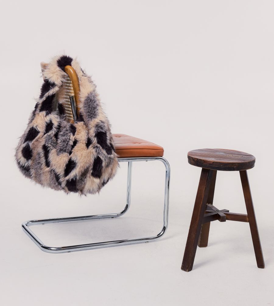 A chunky leopard-print faux fur handbag with a gold-tone handle is draped over a cantilever chair with a tan leather seat, next to a small wooden stool, against a plain light gray background.
