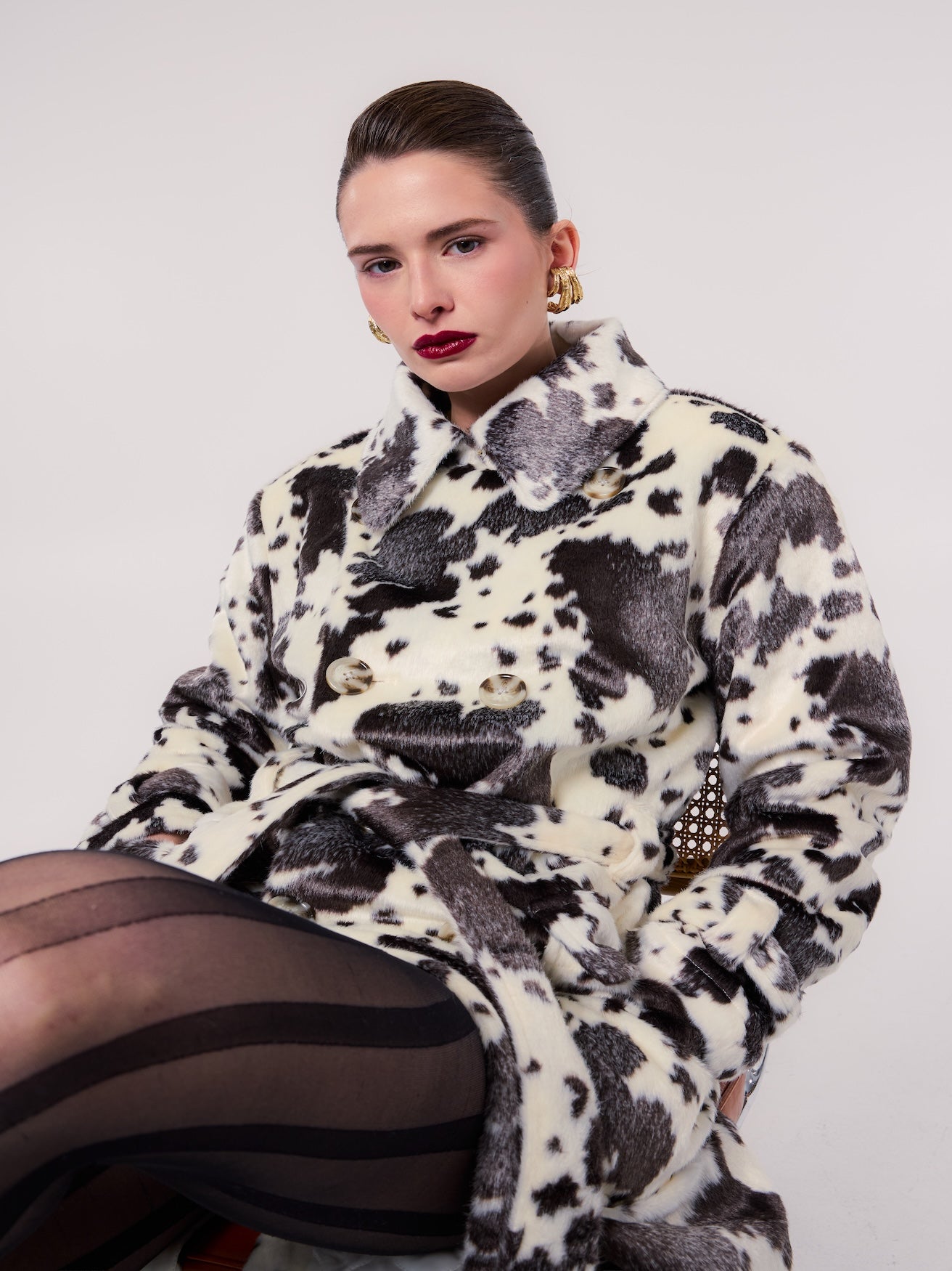 GRACE trench coat in Cow Noir — model wearing a belted vegan faux fur trench coat with a bold black-and-ivory cow print, styled with sheer striped tights and statement gold earrings for a modern editorial look.