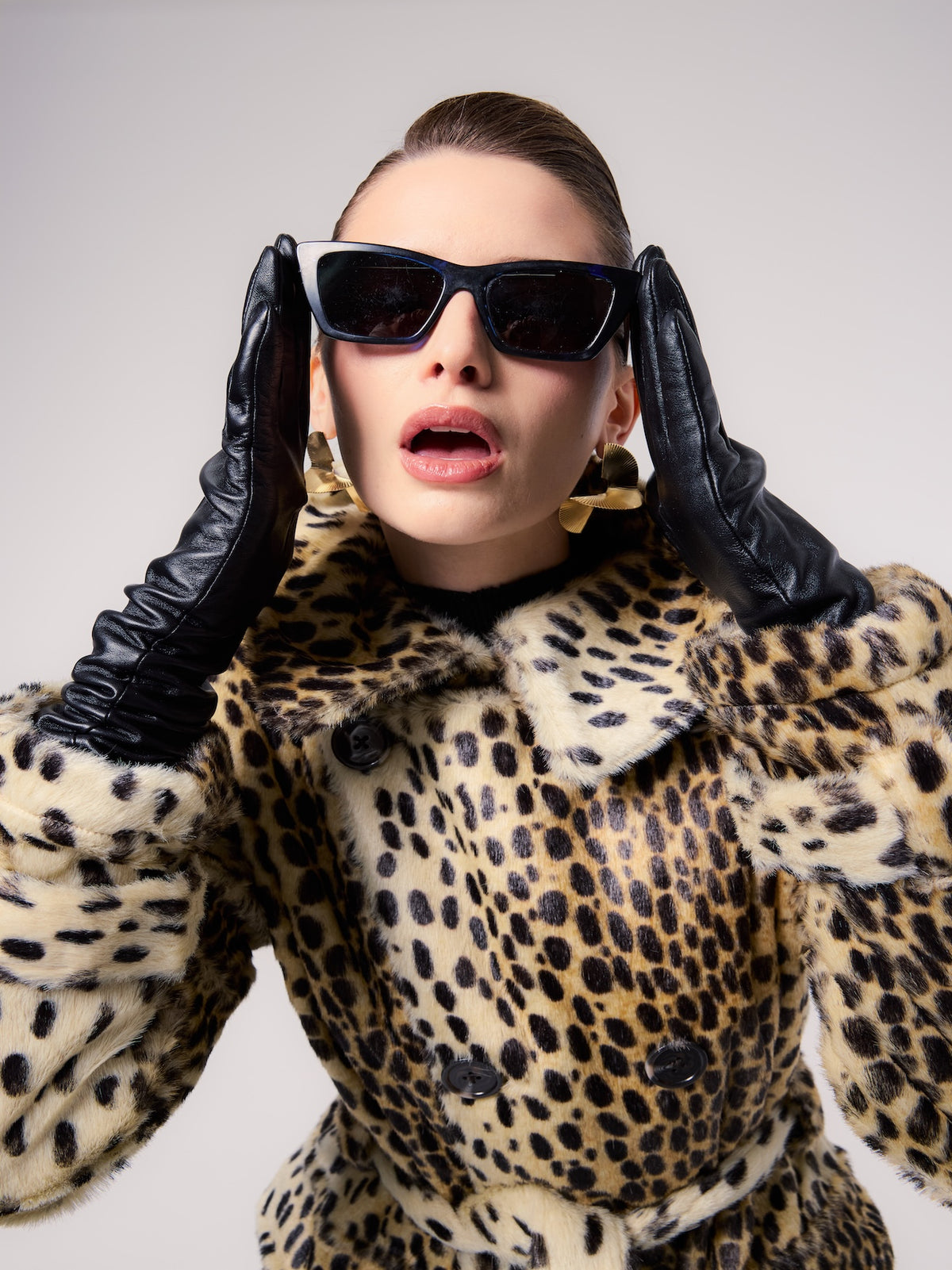 GRACE jacket in Leopard — model wearing a belted vegan faux fur jacket with a classic leopard print, styled with black leather gloves, oversized sunglasses, and statement gold earrings for a bold editorial look.