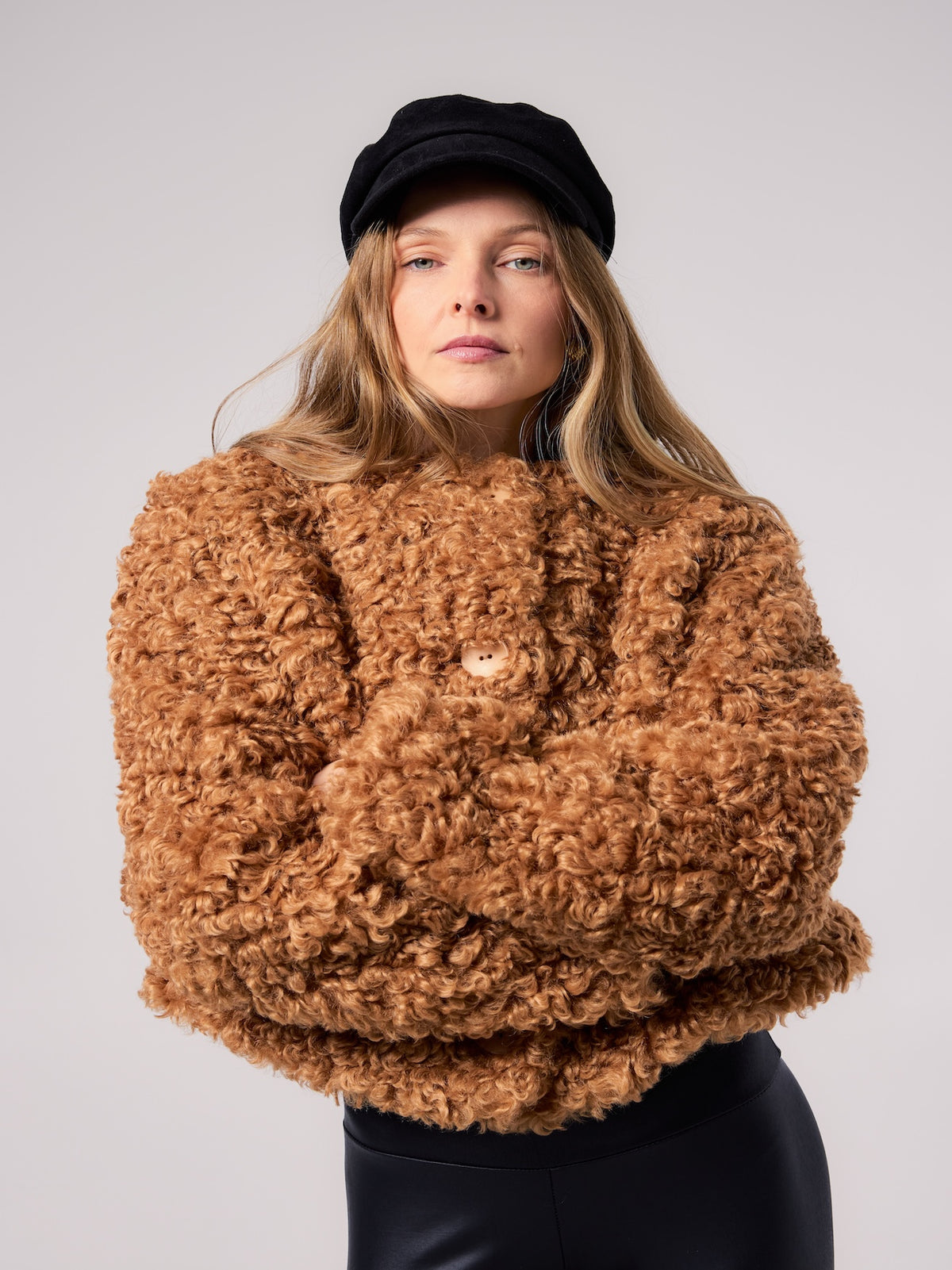 Model wearing a cropped vegan faux fur jacket with a rich curly texture and voluminous silhouette, styled with a black baker boy cap for a modern cold-weather look.
