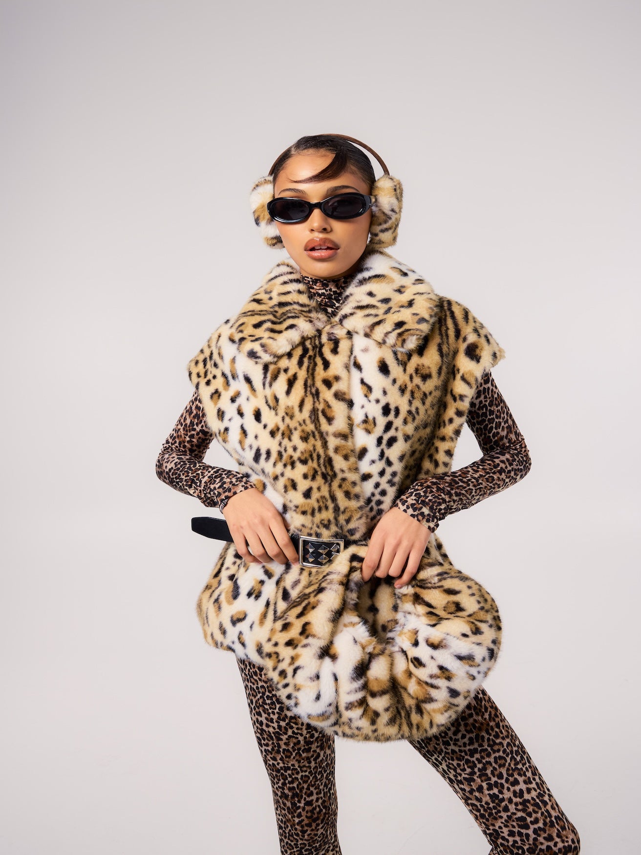 MIA vest in Leopard — model wearing a belted vegan faux fur vest with a bold leopard print, styled with matching leopard-print earmuffs, a fitted animal-print base layer, and slim sunglasses for a statement editorial look.